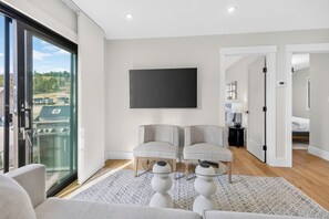 A modern living room with a large TV, two grey armchairs, a glass door leading to a balcony, and a wooden floor.