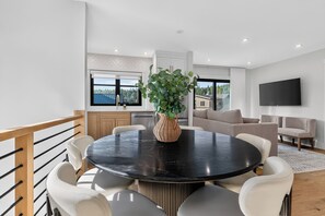 A modern interior with a round dining table, white chairs, a potted plant, a sofa, and a flat-screen TV mounted on the wall.