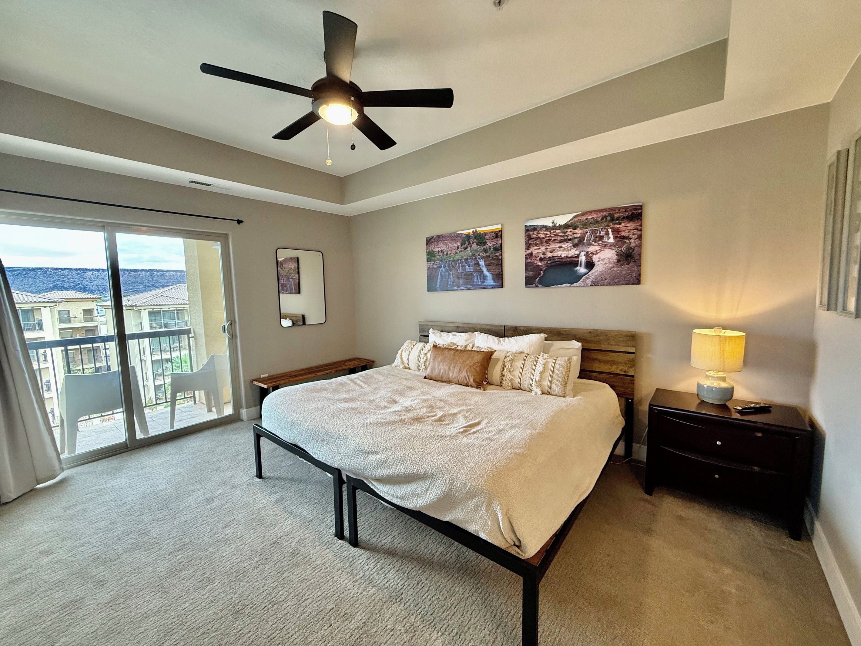 A bedroom with a bed, a nightstand, a lamp, a ceiling fan, a window with a view of buildings, and a wall with three framed pictures.