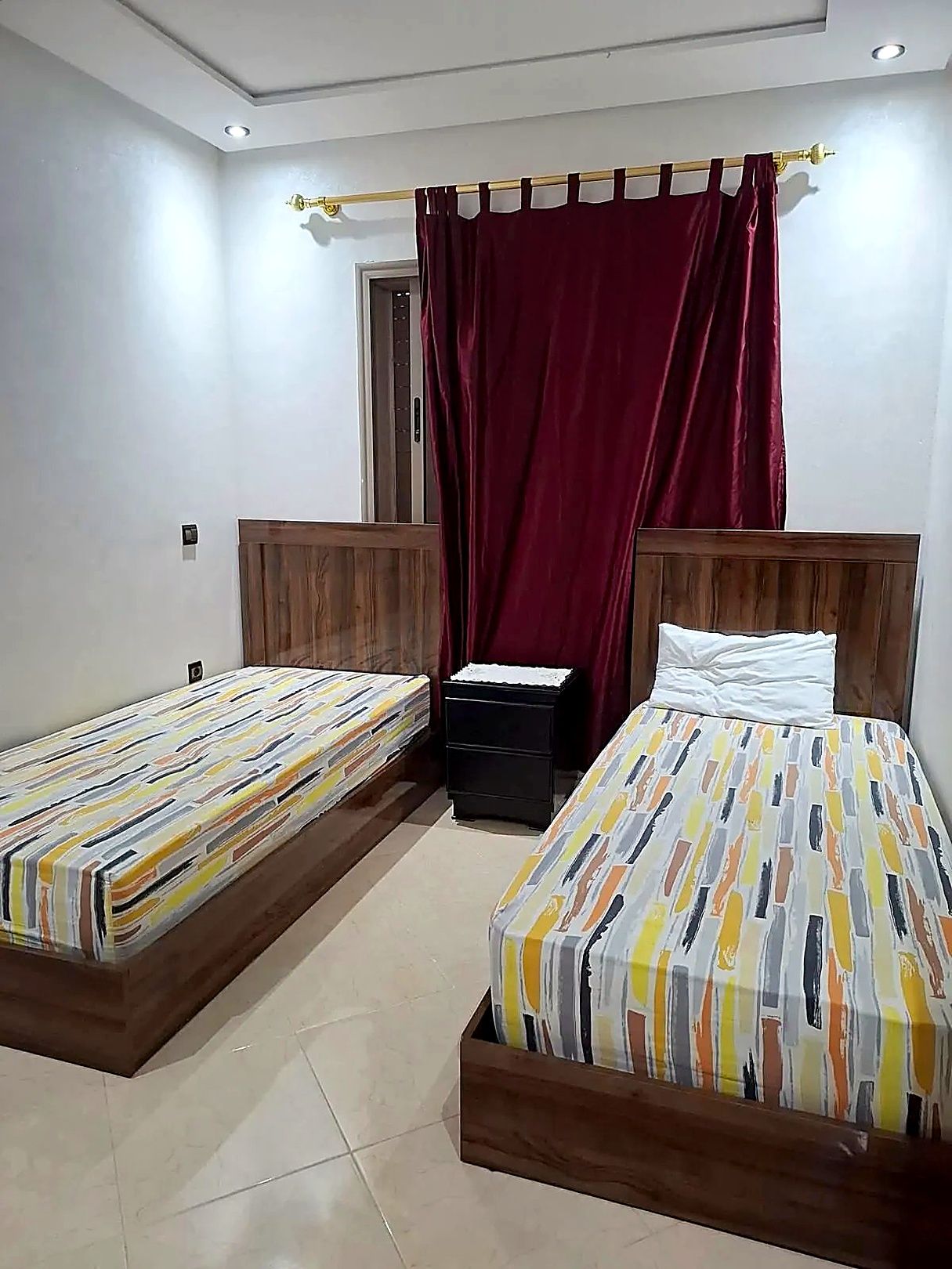 A bedroom with two single beds, a wooden headboard, a small bedside table, and a red curtain.