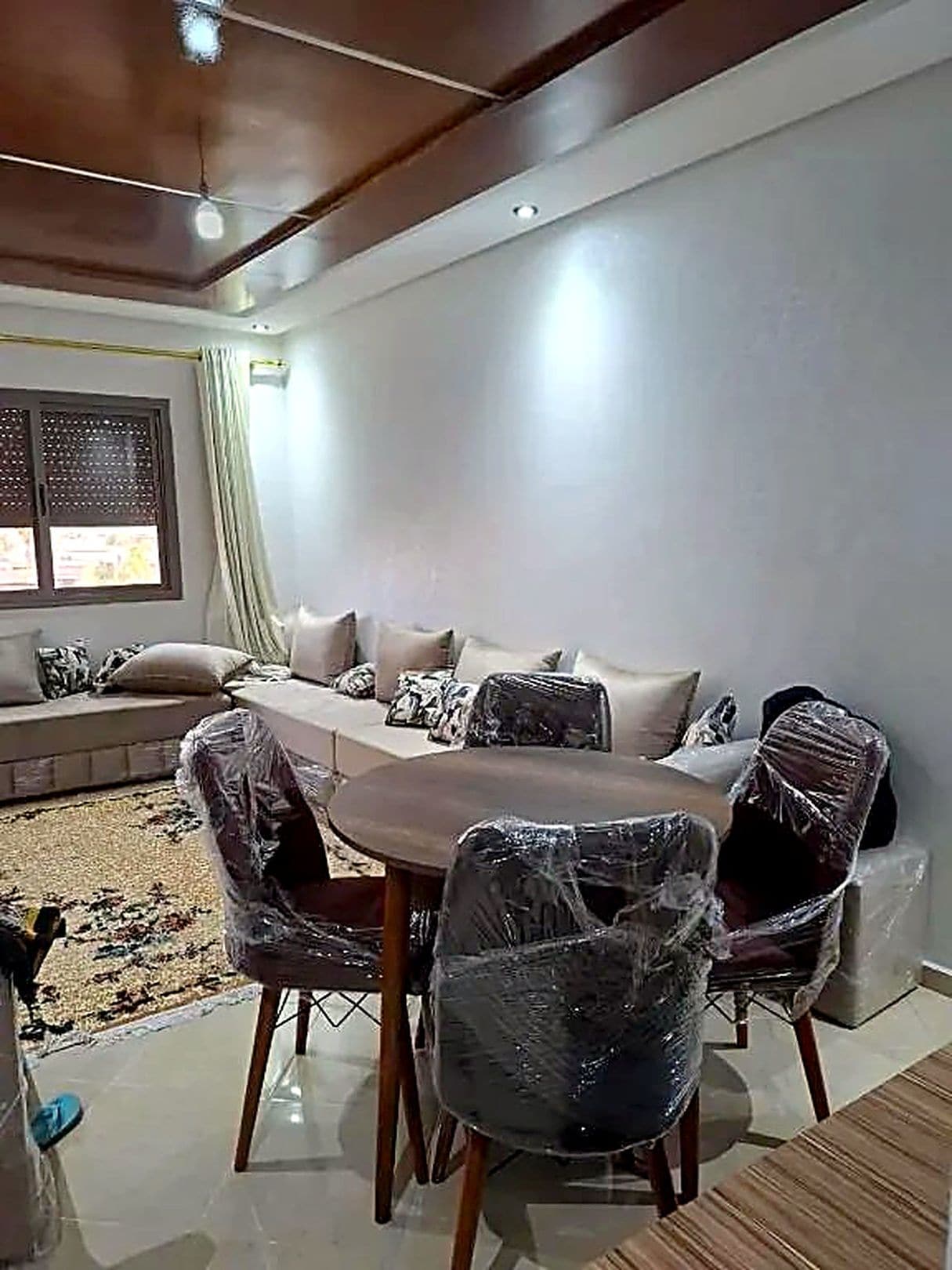 A dining area with a round table and chairs, a sofa with cushions, and a window with blinds.