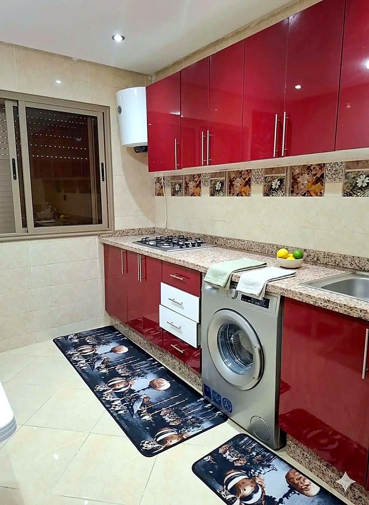 Une cuisine avec des meubles de rangement rouges, une cuisinière à gaz, un lave-linge et une fenêtre avec des stores.