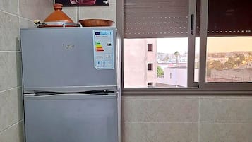 A kitchen with a refrigerator, a window with blinds, and a view of buildings outside.