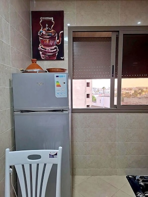 A kitchen with a refrigerator, a window with blinds, and a view of buildings outside. - New Listing (Kénitra)