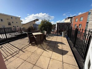 Una terraza en la azotea con una mesa y sillas, rodeada por una cerca de metal negro y edificios al fondo.