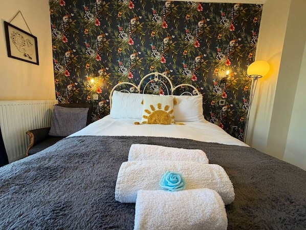 Habitación de hotel con cama, cabecera con diseño de sol, pared con estampado tropical y lámpara en la mesita de noche.