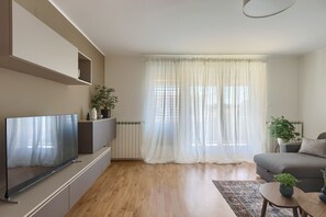 Un salon moderne doté d’un téléviseur à écran plat, d’un canapé gris, d’une table basse et d’une grande fenêtre ornée de rideaux légers.