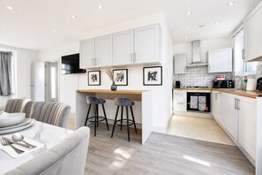 Una cocina moderna con armarios blancos, encimera de madera y horno empotrado. Hay dos taburetes de bar y una mesa de comedor con mantel blanco.