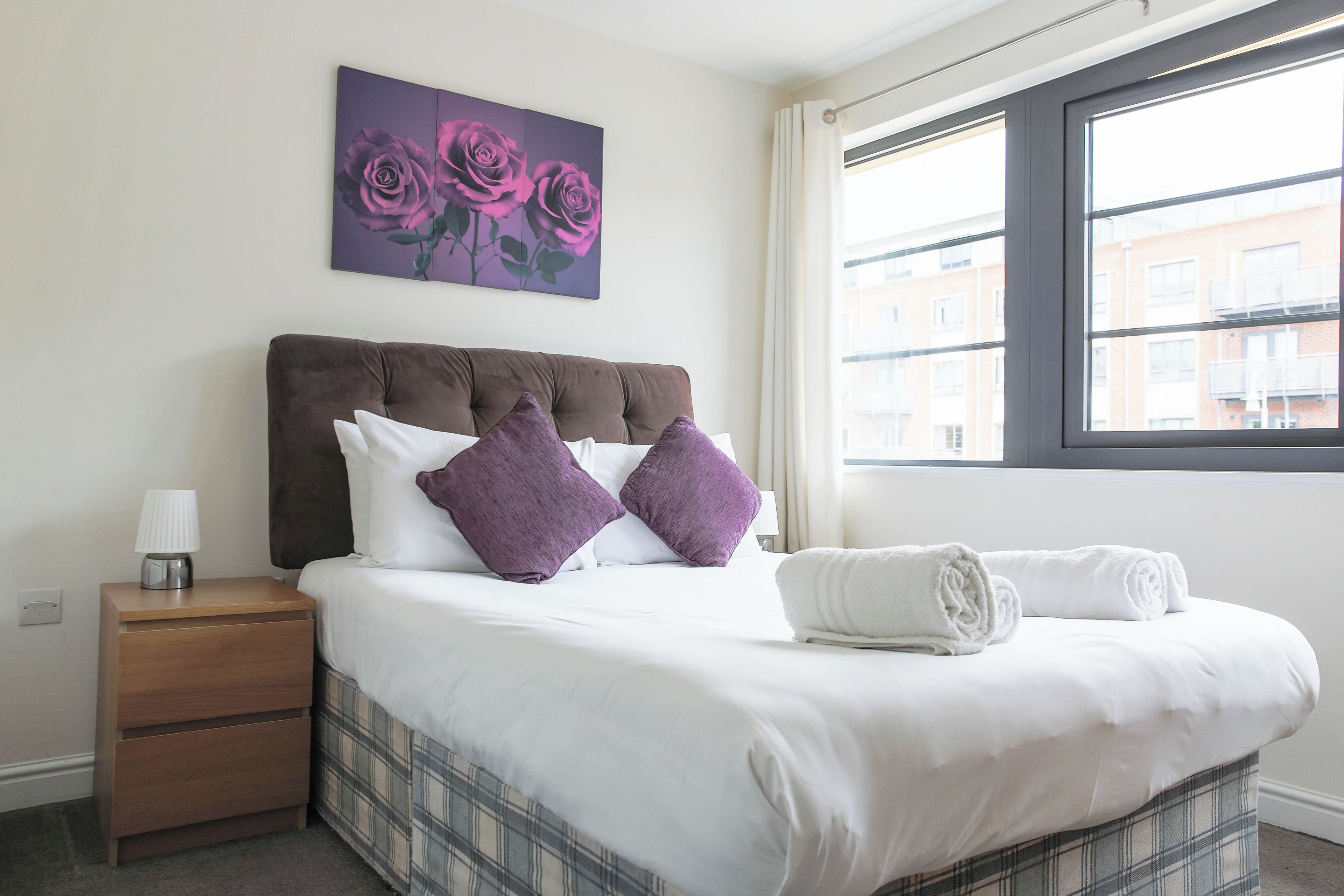 A hotel room with a bed, a nightstand, a lamp, a window with a view of buildings, and a painting of roses.
