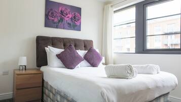 A hotel room with a bed, a nightstand, a lamp, a window with a view of buildings, and a painting of roses.