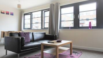 A modern living room with a black leather sofa, a wooden coffee table, and a view of the city through large windows.