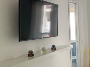 Un téléviseur à écran plat fixé sur un mur blanc, une petite étagère avec des objets décoratifs et une porte donnant sur un balcon.