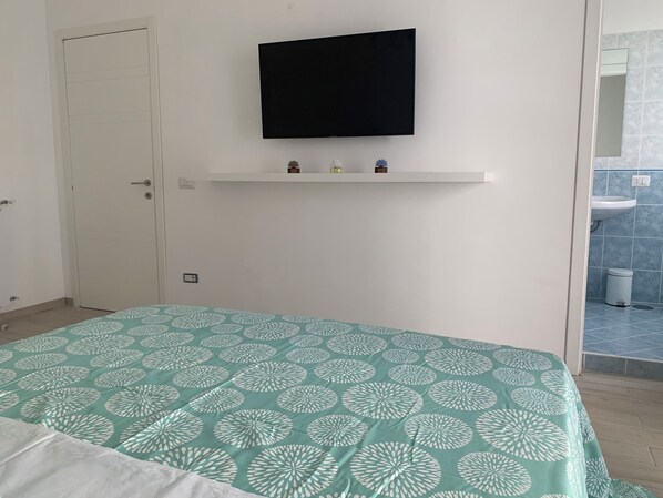 Une chambre à coucher comprenant un lit, un téléviseur fixé au mur, une étagère avec de petits objets, une porte et une salle de bain avec un lavabo et des toilettes.