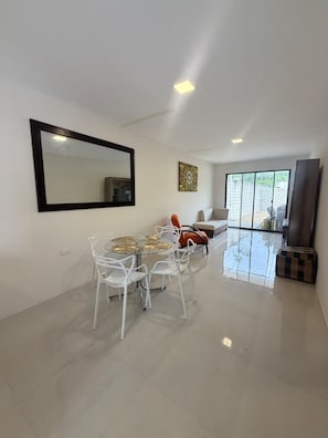 Uma sala de estar ampla com uma mesa de jantar, cadeiras, um sofá, um espelho grande e uma televisão.