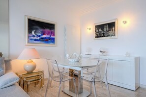 Un comedor moderno con una mesa blanca, sillas transparentes y una mesa de centro de cristal. Hay dos obras de arte enmarcadas en las paredes, un armario blanco con objetos decorativos y una lámpara sobre una mesita auxiliar.