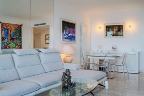 A modern living room with a grey sofa, a glass coffee table, and a dining area with a white table and clear chairs. - New Listing (Cannes)