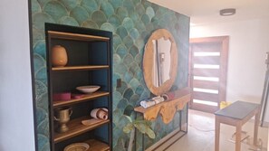 Une pièce comprenant une étagère en bois où sont rangés divers objets, un miroir avec un cadre en bois et une porte dotée de stores horizontaux.