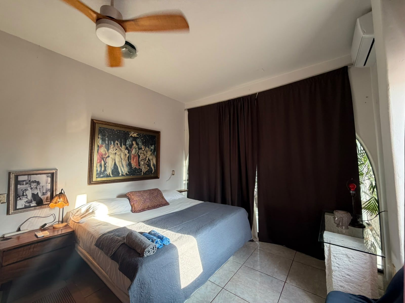 A hotel room with a bed, a ceiling fan, a painting, a nightstand, a lamp, and a window with curtains.