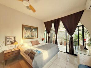 Quarto com cama, mesa de cabeceira, luminária, pintura e amplas janelas com cortinas.