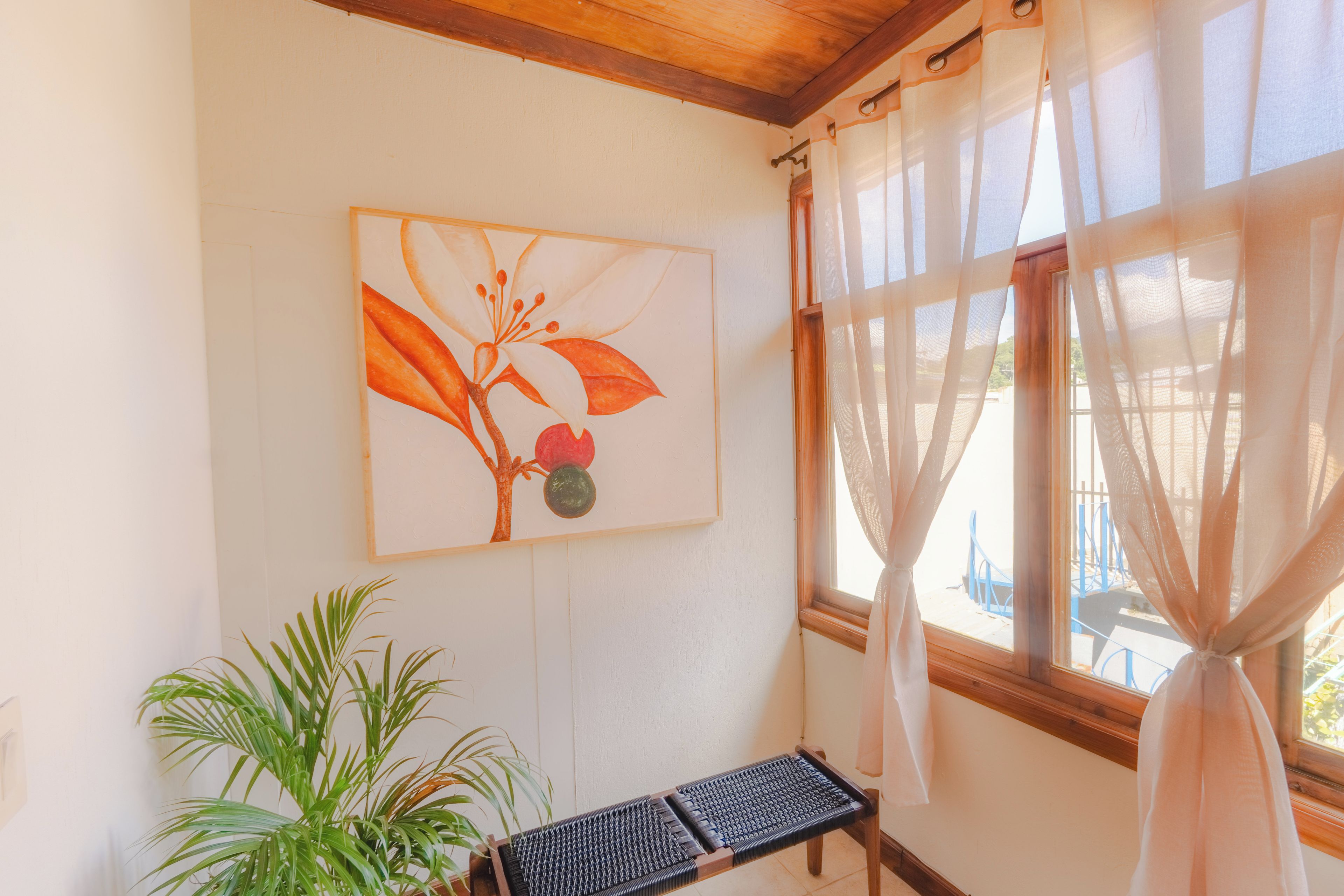 Quarto com teto de madeira, janela com cortinas leves, uma planta e um banco.