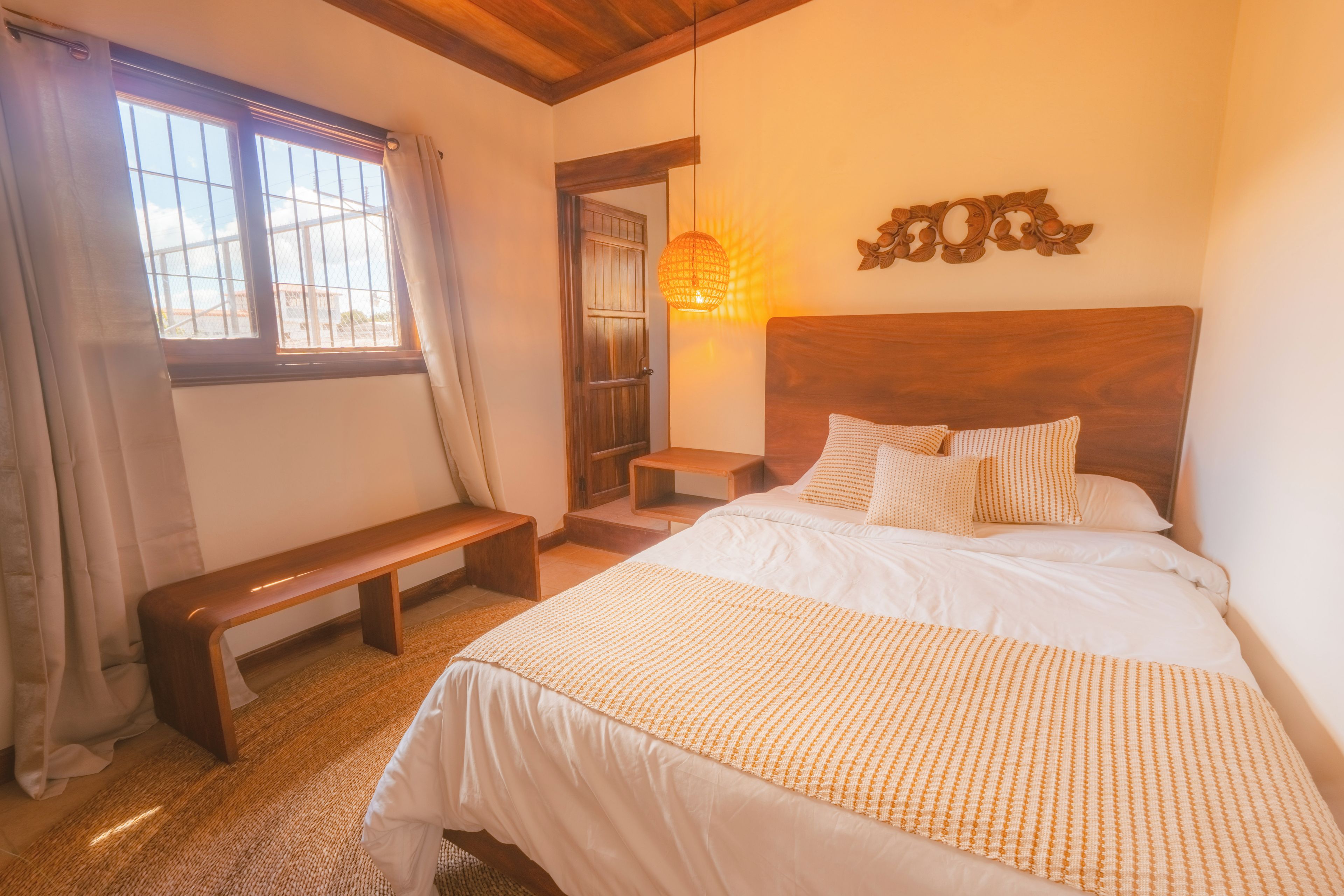 Un dormitorio con una cama de madera, un banco, una ventana con cortinas y una lámpara fijada a la pared.