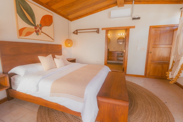 A bedroom with a wooden bed, a nightstand, a wall-mounted lamp, and a door leading to a bathroom. - Flor de café (Juayúa)