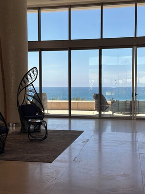 Amplia habitación con ventanas grandes que ofrecen vista al mar, una silla de metal negro y una silla blanca acolchada.