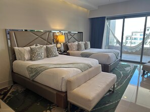 Habitación de hotel con dos camas, un banco, una mesita de noche con lámpara y vistas al exterior.