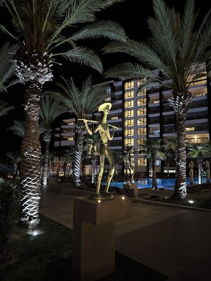 Escena nocturna con una estatua, palmeras, una piscina y un edificio con ventanas iluminadas.