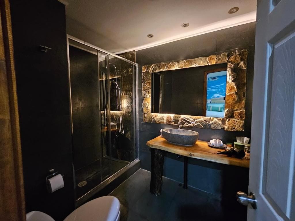 A modern bathroom with a glass-enclosed shower, a stone accent wall, and a wooden vanity with a bowl sink.