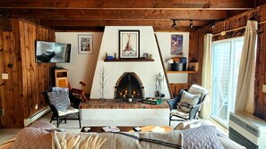 Ein gemütliches Wohnzimmer mit Kamin, Holzwänden, einem Fernseher und einem Fenster mit Vorhängen.