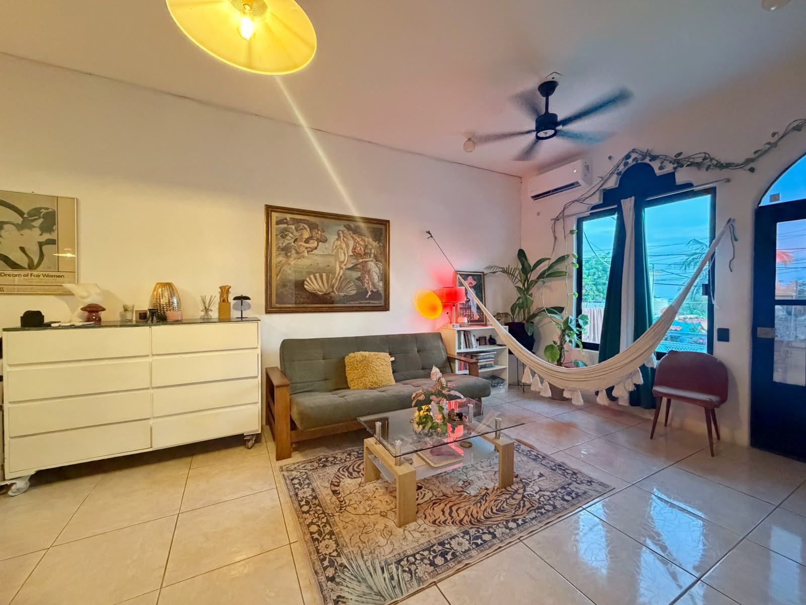 A living room with a sofa, a coffee table, a hammock, a ceiling fan, and a large window.