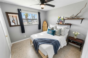 Une chambre avec un lit, un ventilateur de plafond, une planche de surf, une fenêtre avec des rideaux et une étagère fixée au mur, ornée d’objets décoratifs.