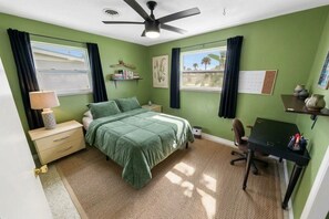 Een slaapkamer met een bed, bureau, stoel, plafondventilator en raam met gordijnen.