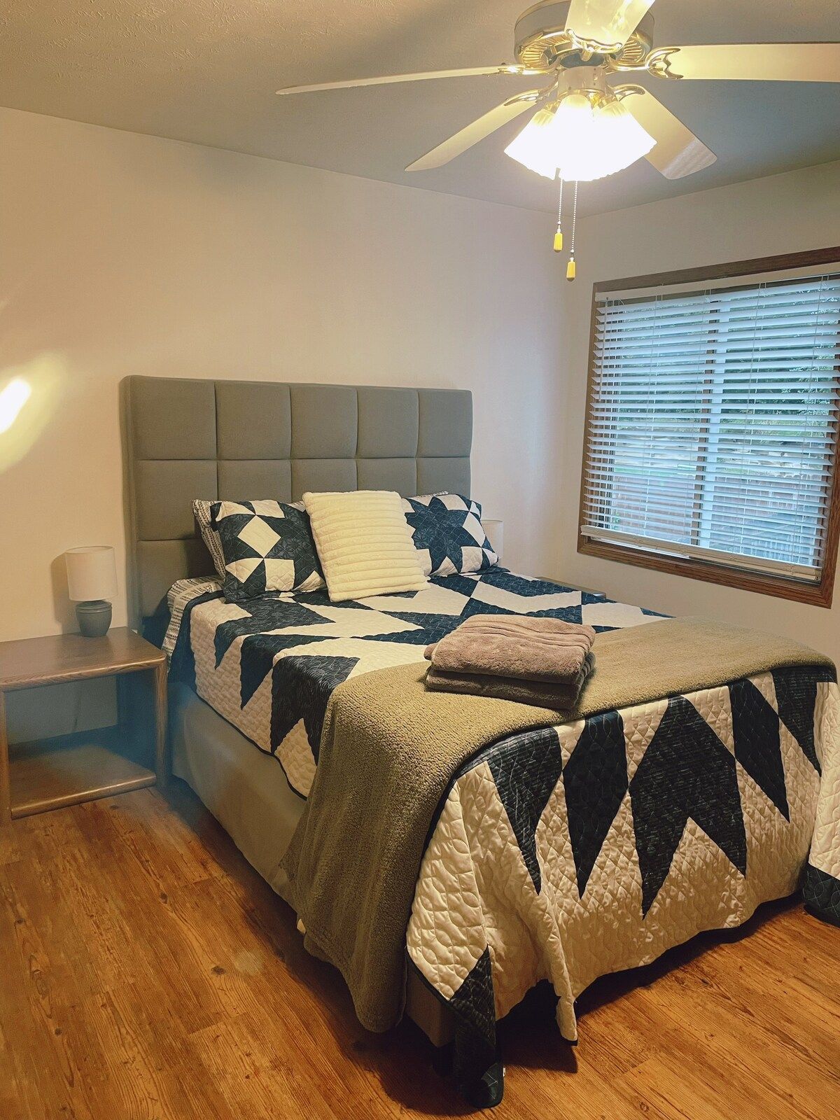 A bedroom with a bed, a nightstand, a ceiling fan, and a window with blinds.
