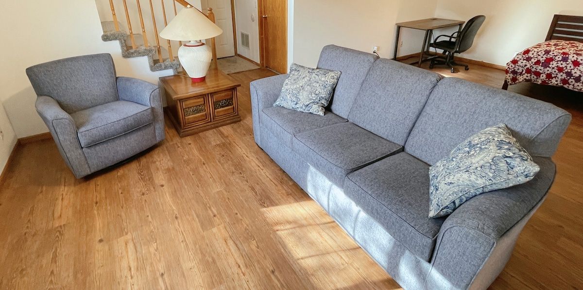A living room with a grey sofa, a matching armchair, a wooden side table with a lamp, and a bed with a floral bedspread.