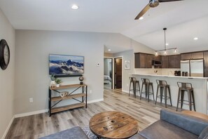 A modern kitchen with bar stools, a dining area with a round wooden table, and a living room with a sofa and a TV mounted on the wall. - Up North Living - Fully furnished updated condo for extended stays in NW WI. (Cumberland)