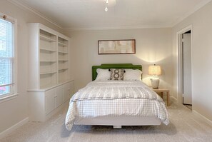 Et soveværelse med seng, natbord, lampe, et vindue med rullegardiner og indbyggede hylder.