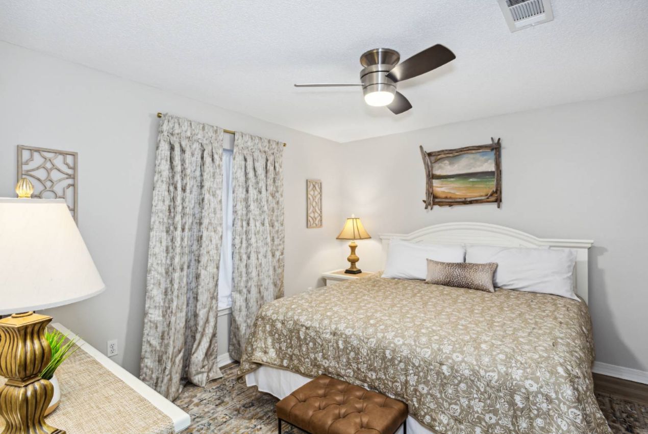 A bedroom with a bed, a nightstand, a lamp, a ceiling fan, and a window with curtains.