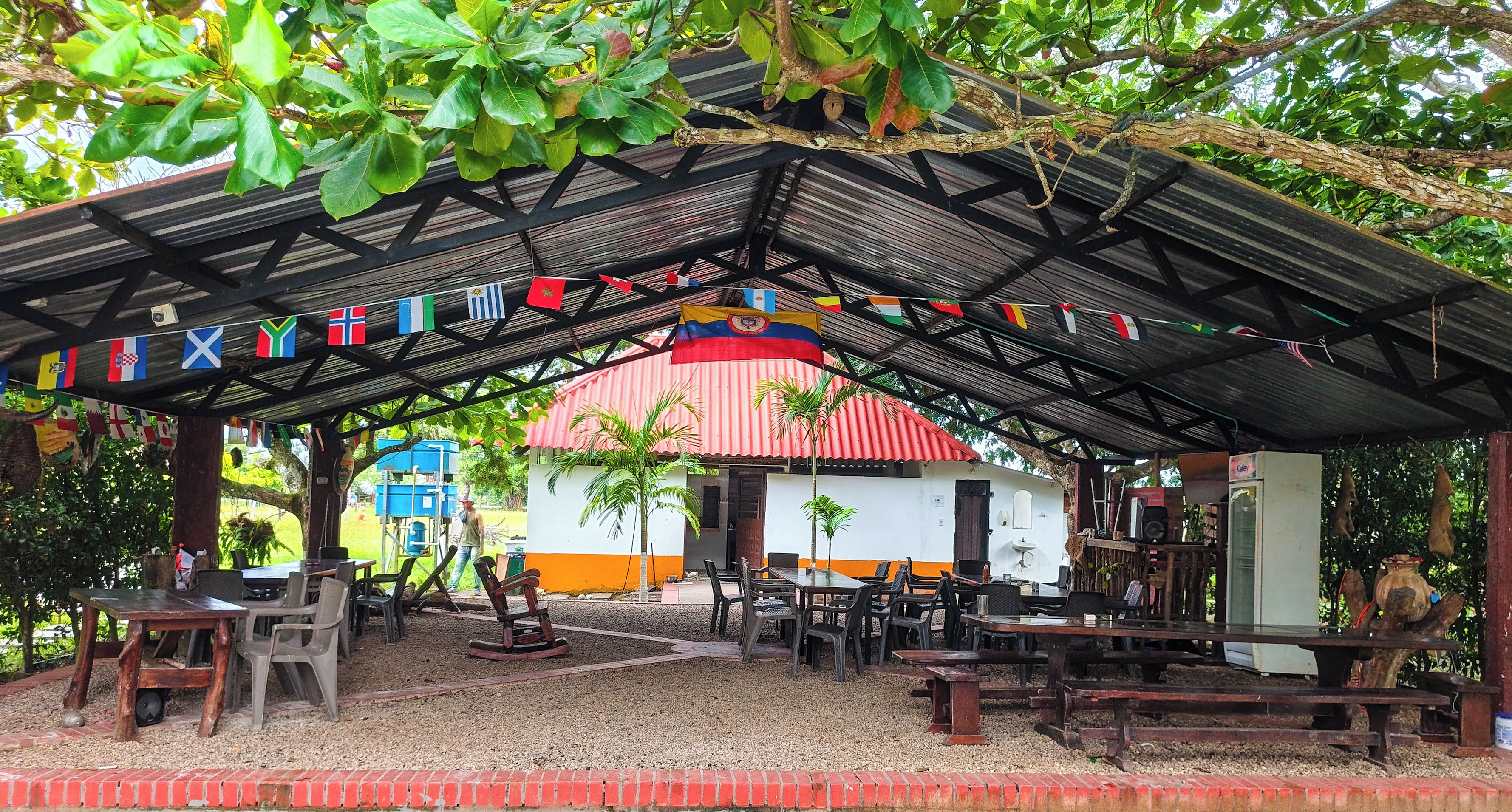 An outdoor seating area with tables and chairs, a covered pavilion, and a building with a red roof in the background.