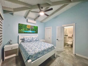 Une chambre à coucher comprenant un lit, une table de chevet, un ventilateur de plafond, un tableau, une porte donnant sur une salle de bain et un sol carrelé.