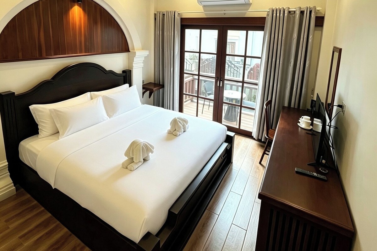 Une chambre d’hôtel avec un grand lit, un plafond en bois, un appareil de climatisation, un bureau avec un ordinateur et une vue sur l’extérieur, visible à travers une porte vitrée.