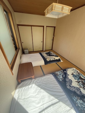 Ein Zimmer mit zwei Betten, einer kleinen Holzbank, einer traditionellen japanischen Schiebetür und einer hängenden Leuchte.