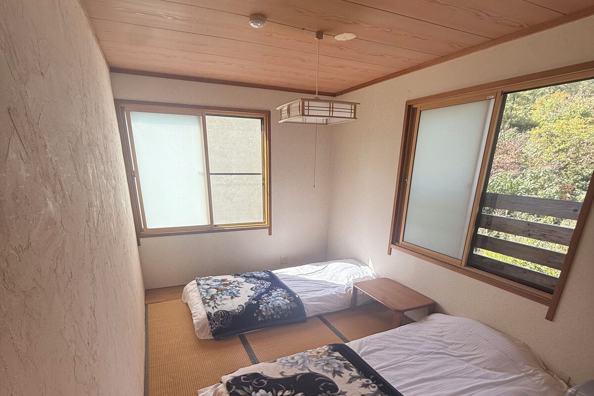 Ein Zimmer mit zwei Betten, Tatami-Matten, einem Deckenventilator und Fenstern mit Holzrahmen.