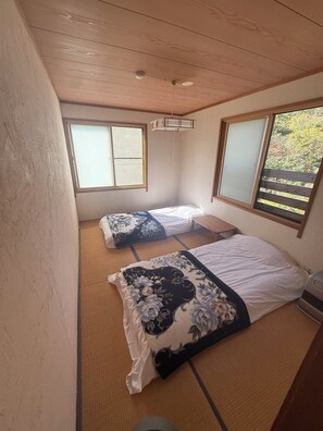 Ein Zimmer mit zwei Betten, Tatami-Matten, einem Deckenventilator und Fenstern mit Holzrahmen.