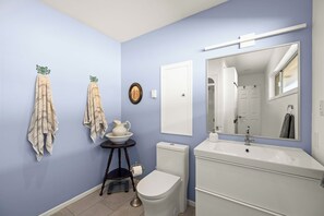 Une salle de bain avec des toilettes, un lavabo et un miroir. Des serviettes sont accrochées au mur, et une petite table est posée là, avec une carafe et un bol.