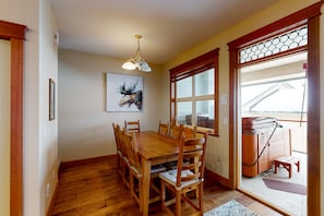 A dining area with a wooden table and chairs, a mounted artwork, and a view of a hot tub outside. - Tree Tops by Okanagan Premier (Beaverdell)