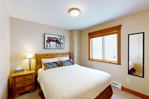 A bedroom with a bed, a nightstand with a lamp, a window, a wall-mounted mirror, and a painting of a deer. - Tree Tops by Okanagan Premier (Beaverdell)