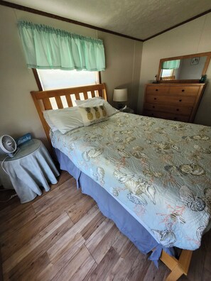Une chambre à coucher comprenant un lit, une commode, une table de chevet avec une lampe, une petite table avec un ventilateur et une fenêtre avec un rideau.