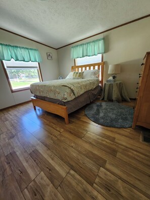Une chambre à coucher avec un lit en bois, une table de chevet avec une lampe, un petit tapis rond et une fenêtre avec des rideaux.